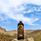 A dark bottle labeled 'Muddy Moose' stands on a rock in a desert landscape with mountains and a blue sky with clouds in the background.