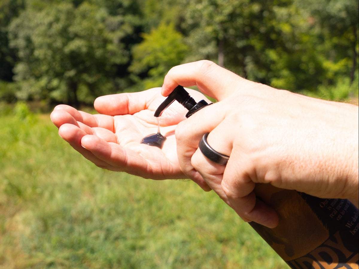 A close-up of a person's hands dispensing a dark liquid from a pump bottle onto their palm, with a blurred outdoor background of green trees and grass.